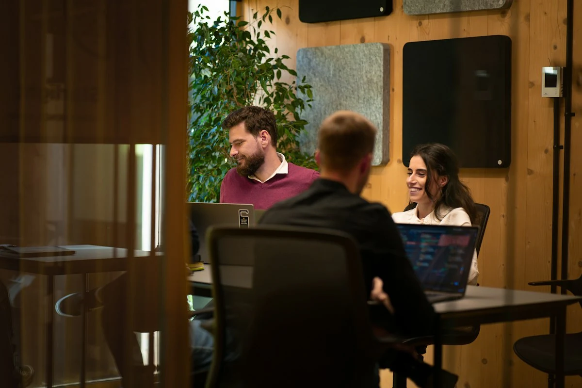 Trois personnes souriantes travaillant sur des ordinateurs portables autour d'une table, dans une salle aux murs en bois habillés de panneaux acoustiques