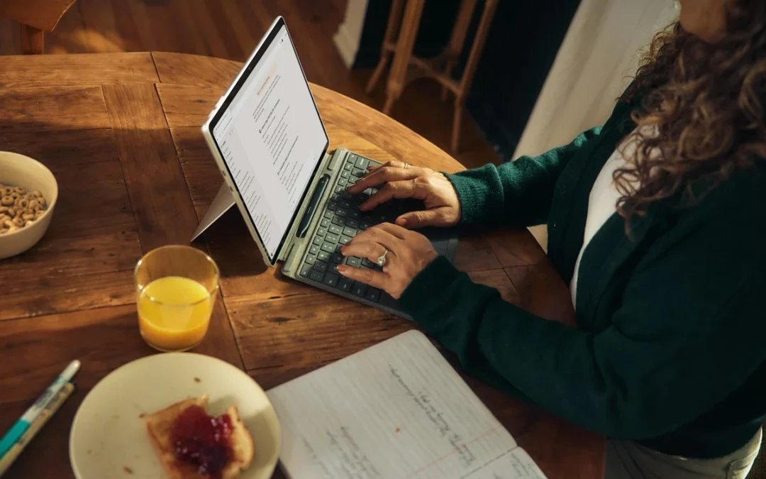 Femme en gilet vert tapant sur un ordinateur portable posé sur une table en bois, à côté d'un jus d'orange, d'une tartine et d'un cahier ouvert